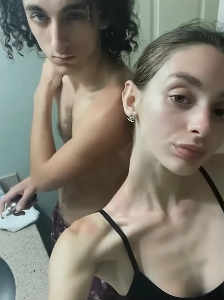 A young man with dark curly hair and a young woman with light hair take a selfie in a bathroom.