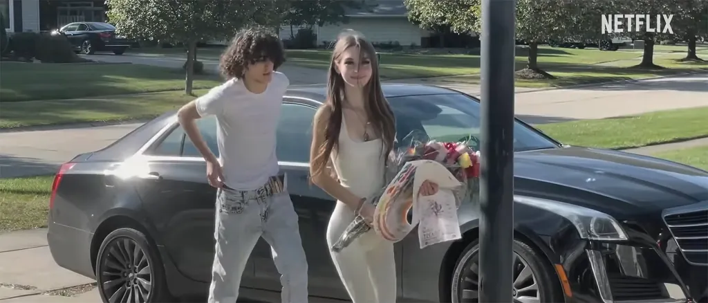 A teenage girl in a cream dress holds flowers and a colorful bag next to a boy in white jeans as they stand by a dark car.