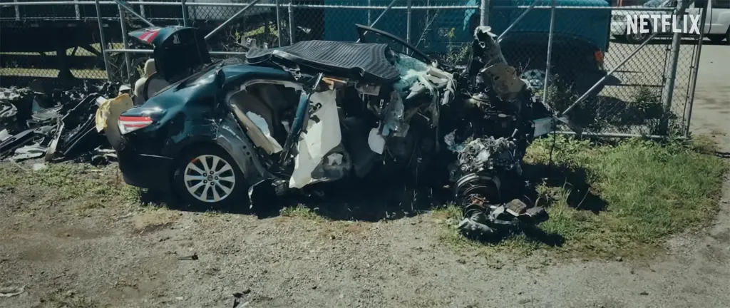 A completely demolished car sits beside a chain-link fence, with its back half largely intact but the front half reduced to unrecognizable debris.