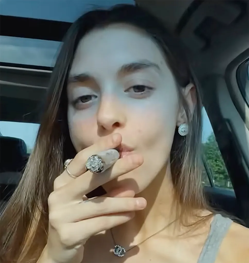 A young woman with light skin and dark hair sits in a car, exhaling smoke from a cigarette.