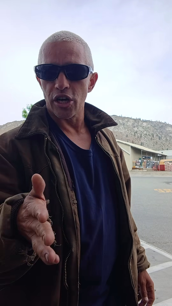 A man wearing sunglasses, a dark blue shirt, and a brown jacket, extends his right hand forward while speaking, with a mountain and building in the background.