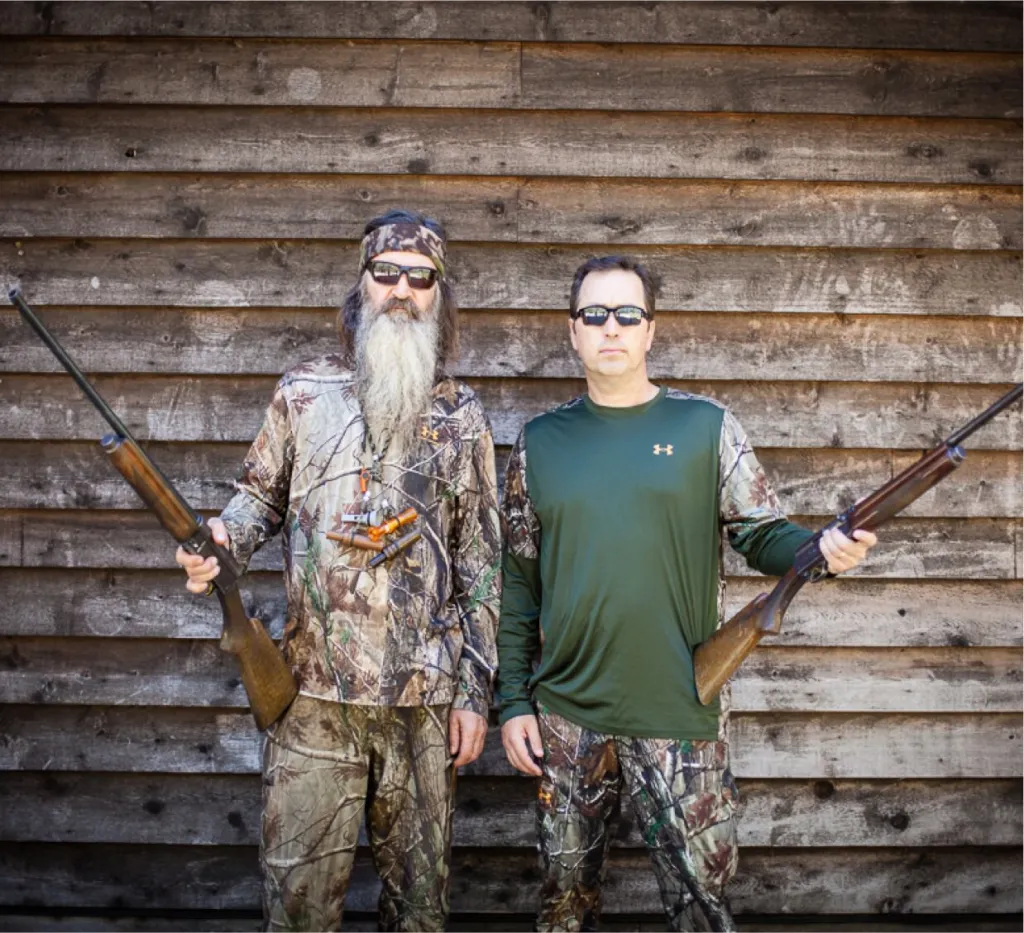 Two men in camouflage, holding rifles, stand in front of a wooden wall.