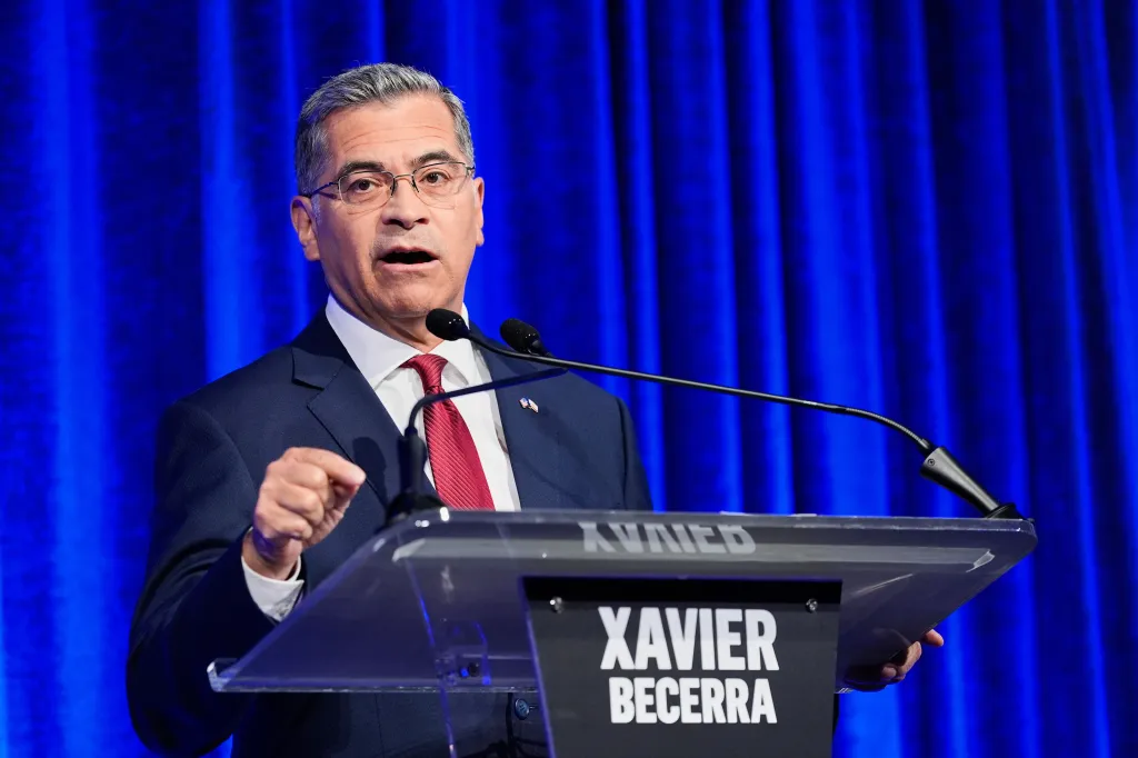 Xavier Becerra speaking at a podium with a microphone, against a blue curtain backdrop.