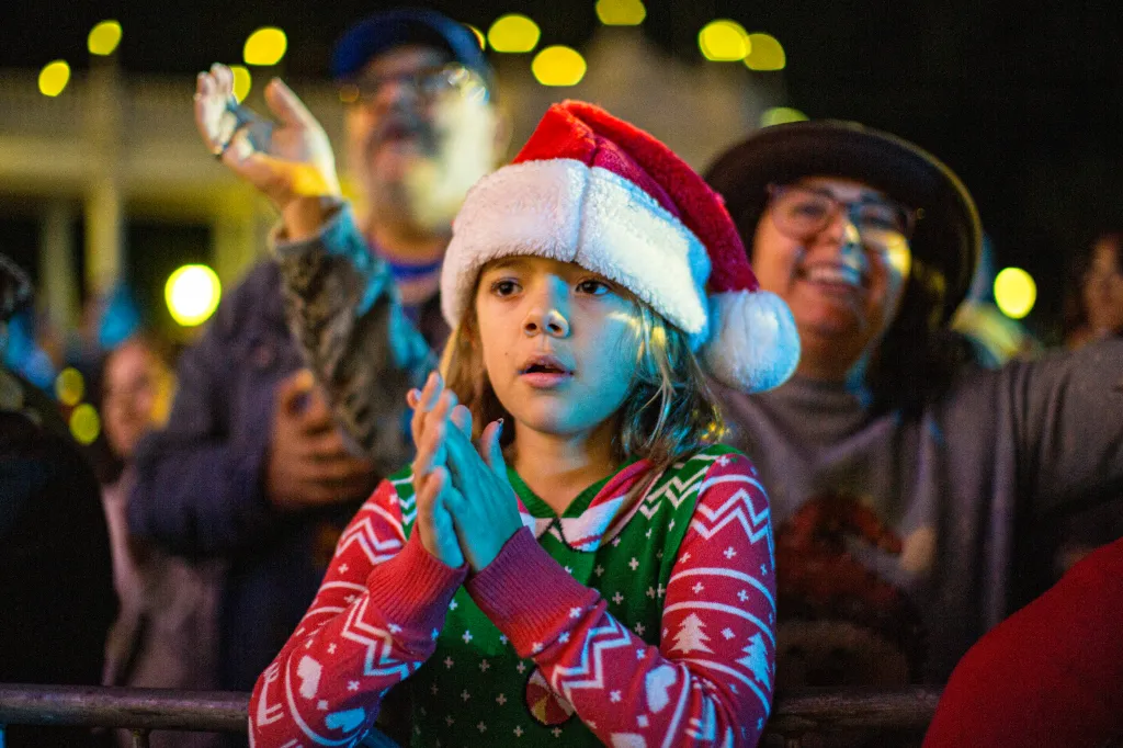 A young fan in a Santa hat and a festive sweater watching a performance.