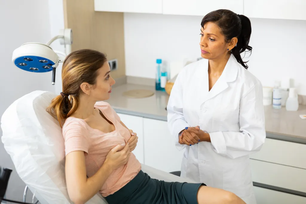 A woman in a peach shirt discusses breast augmentation with a female doctor.