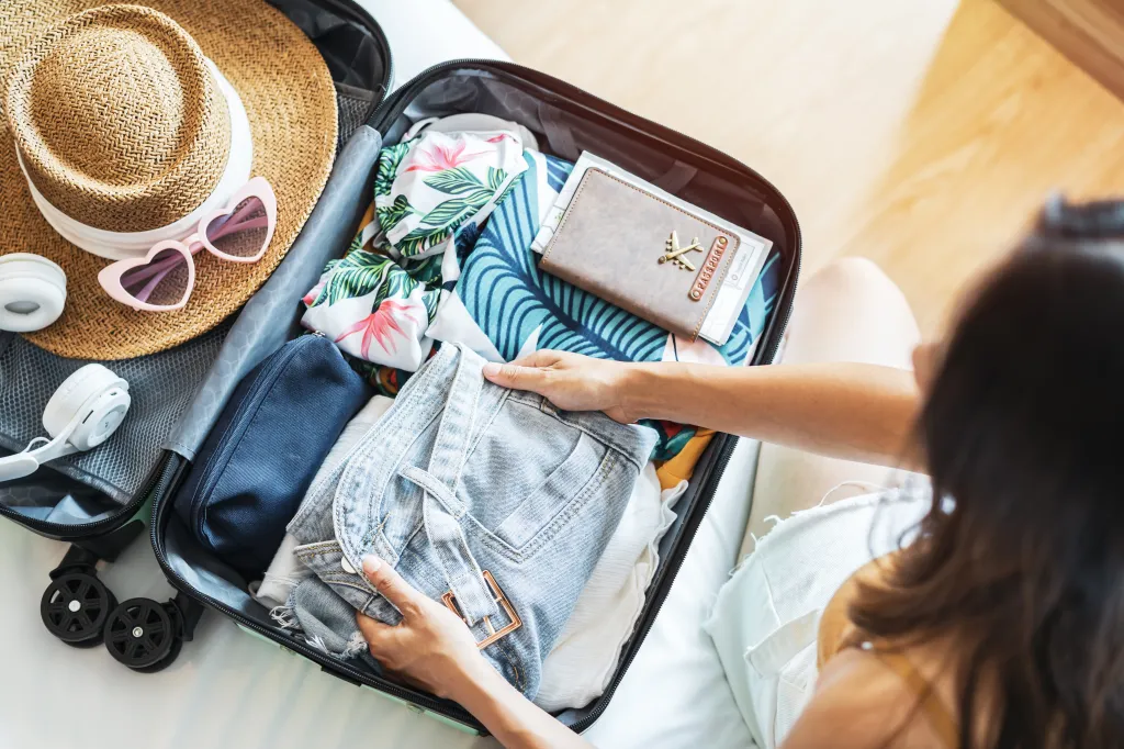 A woman packing a suitcase with clothes, a straw hat, sunglasses, headphones, and a passport holder.