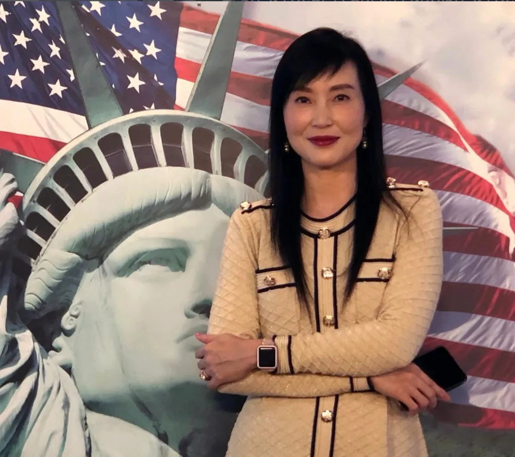 Yvonne Yiu, a woman with long dark hair, stands in front of a depiction of the Statue of Liberty and the American flag.