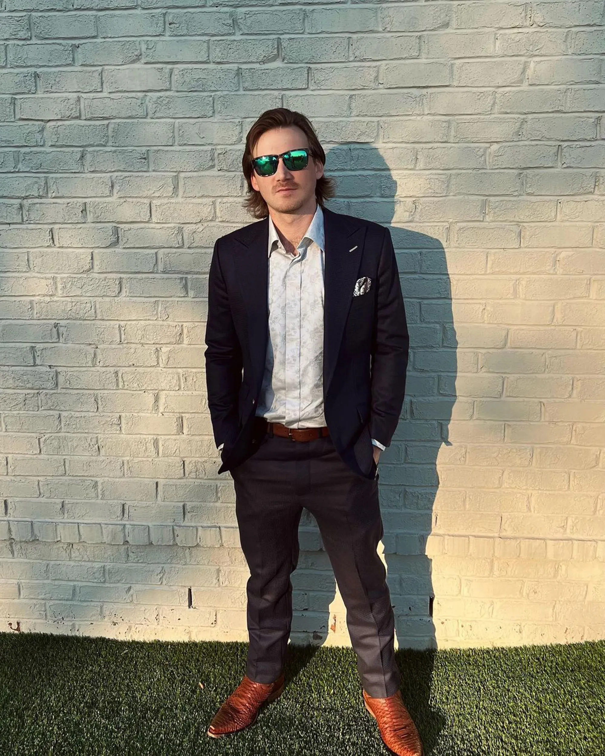 Morgan Wallen wearing a suit, button-down shirt, sunglasses, and cowboy boots, standing in front of a white brick wall.