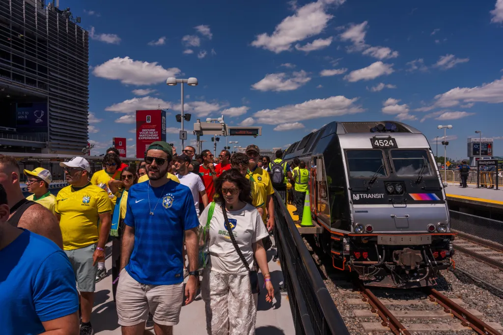 Fans in Brazil and Morocco jerseys walk past an NJ Transit train at Meadowlands station, heading towards MetLife Stadium for a World Cup match.