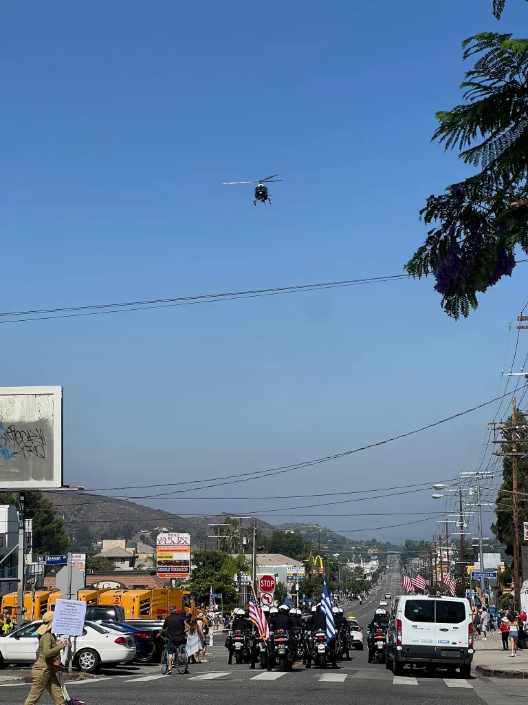 A helicopter flies over a street scene with motorcycles, a van, and people, with hills in the background.
