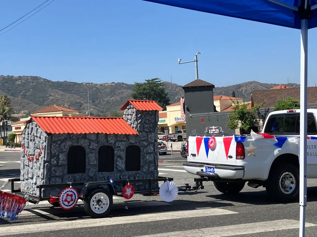 A pickup truck decorated with red, white, and blue streamers and stars, pulling a trailer with a grey, stone-like structure with a red roof, in front of a mountainous background.