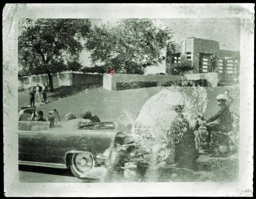 Black and white photo of a dark vehicle with people inside, traveling on a road, with a man on a motorcycle escorting it.