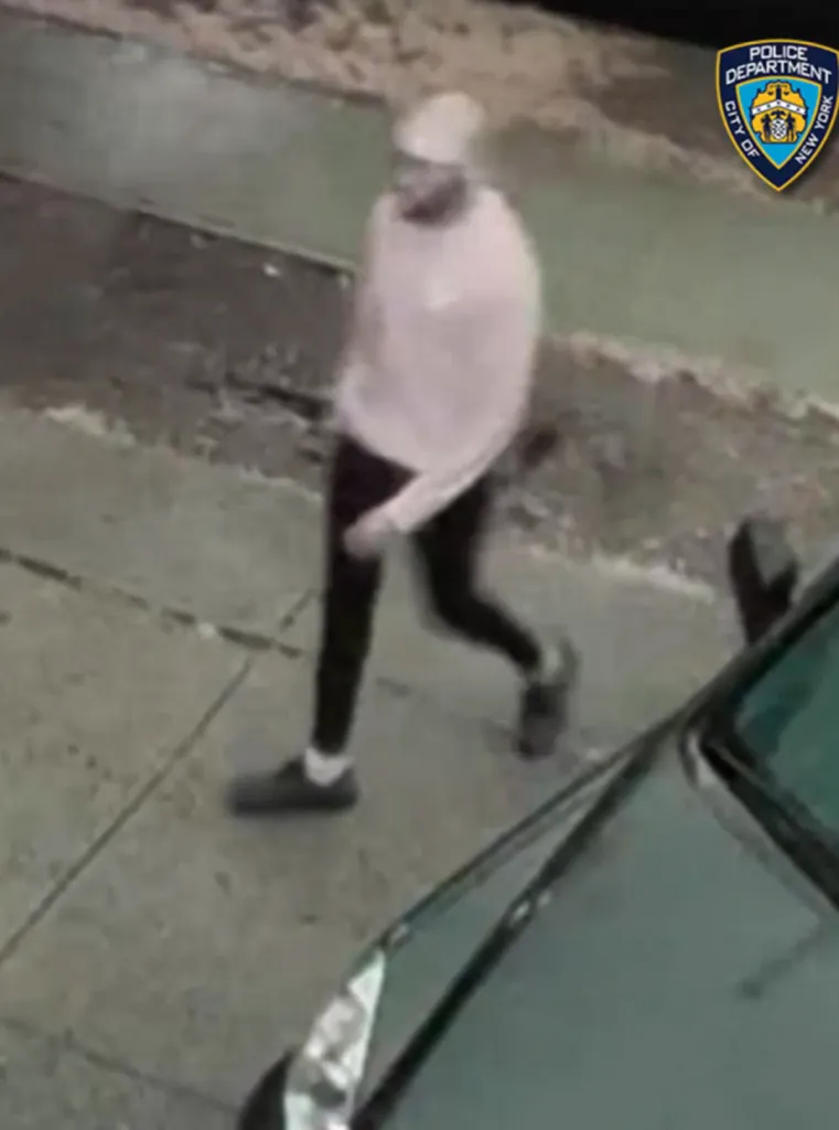 Surveillance footage of an individual in a light-colored sweatshirt and dark pants walking on a sidewalk next to a car.