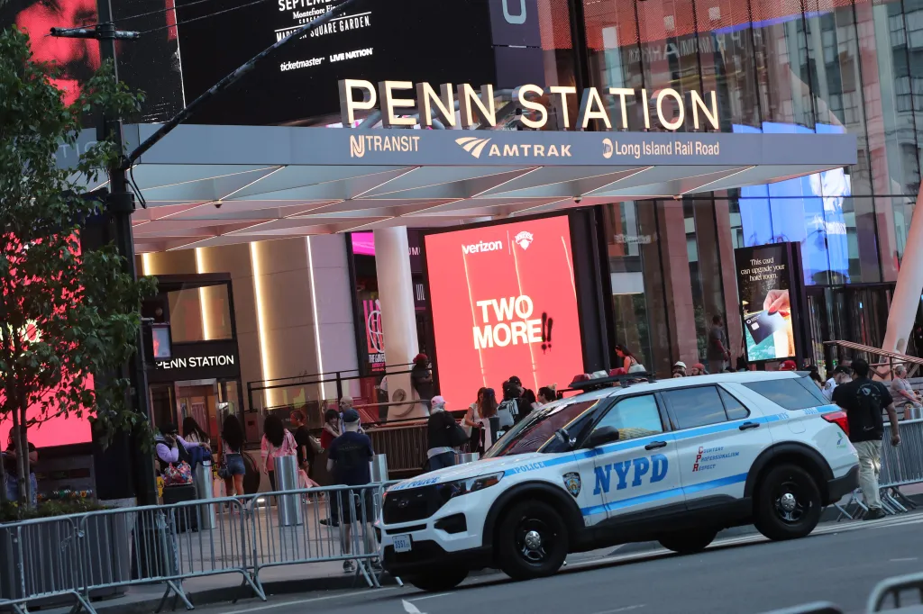 A white NYPD police SUV parked on a street outside Penn Station in NYC, with pedestrians and large video billboards visible.