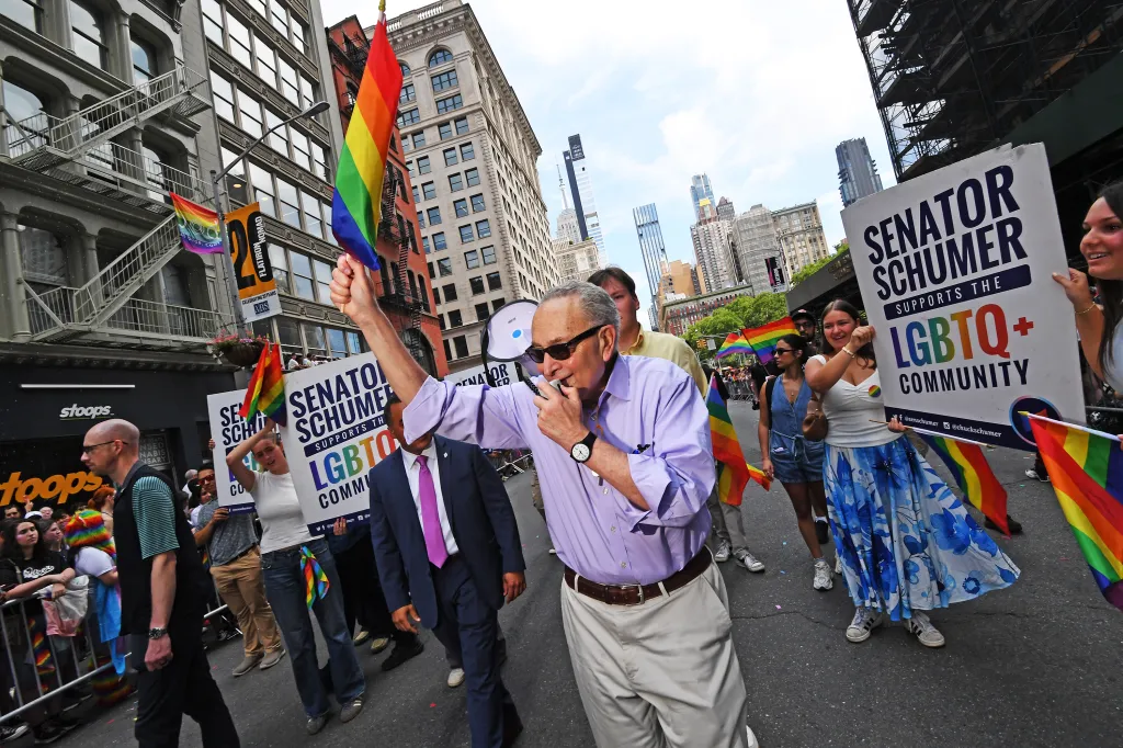 Senator Schumer addressing the crowd at a Pride parade, surrounded by supporters holding rainbow flags and signs.