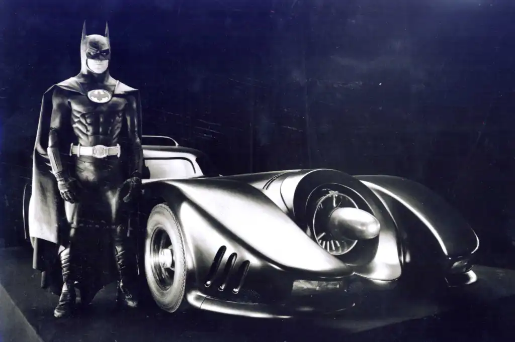 Michael Keaton as Batman standing next to the Batmobile from the movie