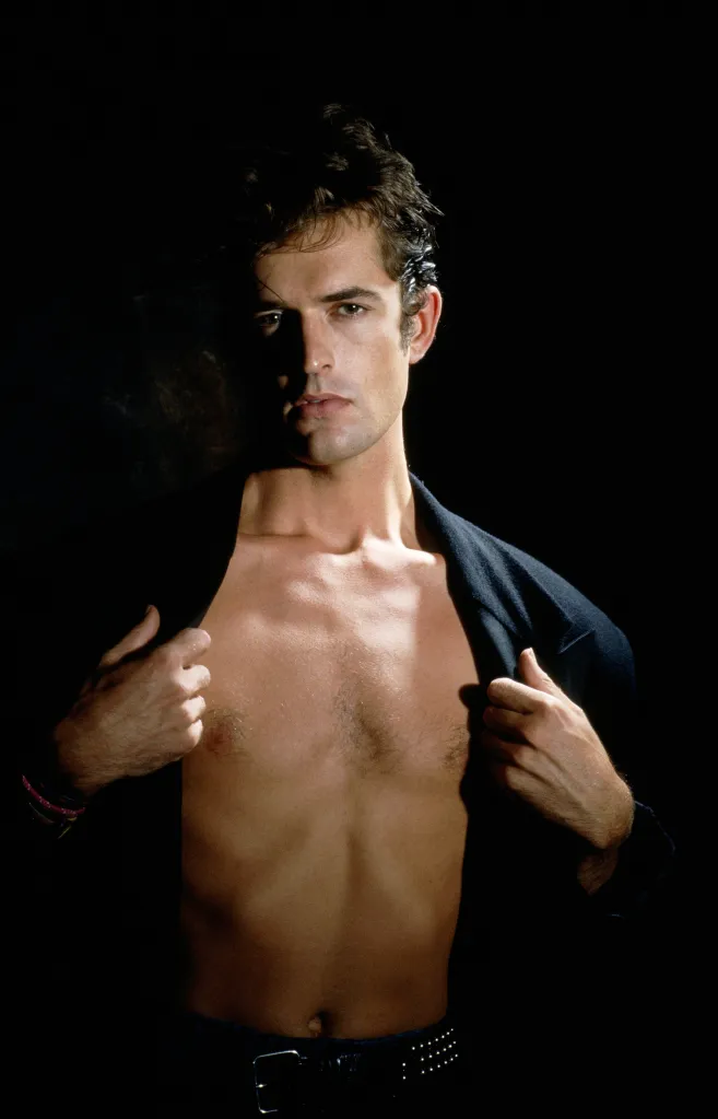 British actor Rupert Everett posing shirtless with a jacket over his shoulders.