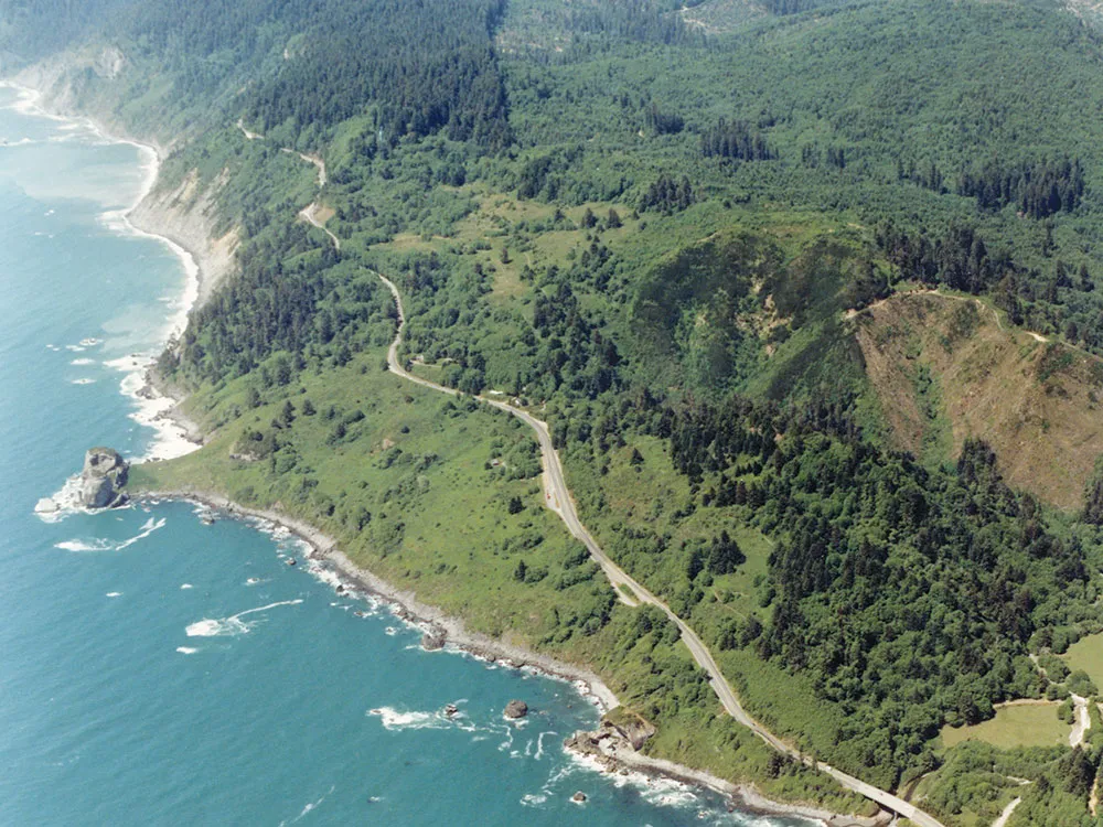 Aerial view of US Highway 101 winding along the Del Norte County coastline, with a forested hillside and the Pacific Ocean.