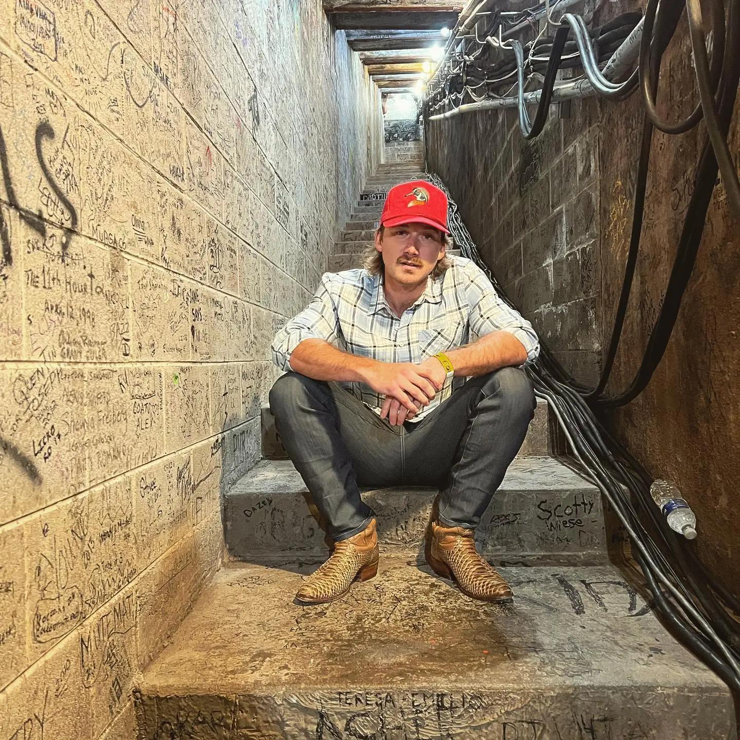 Morgan Wallen in a red hat, plaid shirt, and cowboy boots sits on concrete stairs with graffiti-covered walls.