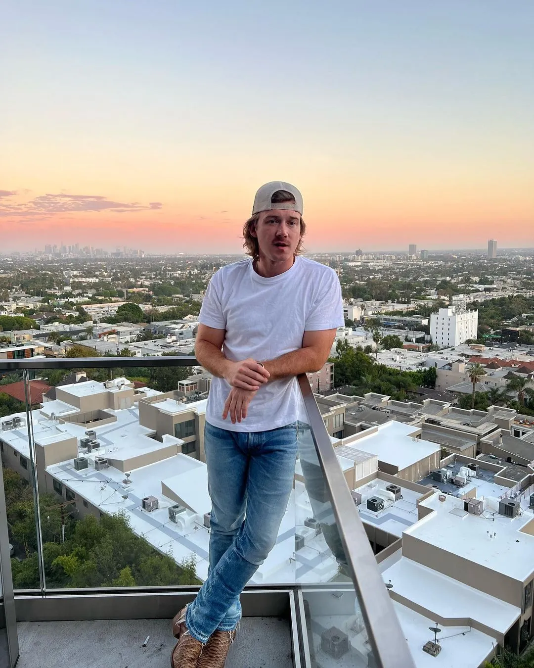 Man in a white t-shirt and jeans standing on a balcony overlooking a city at sunset.