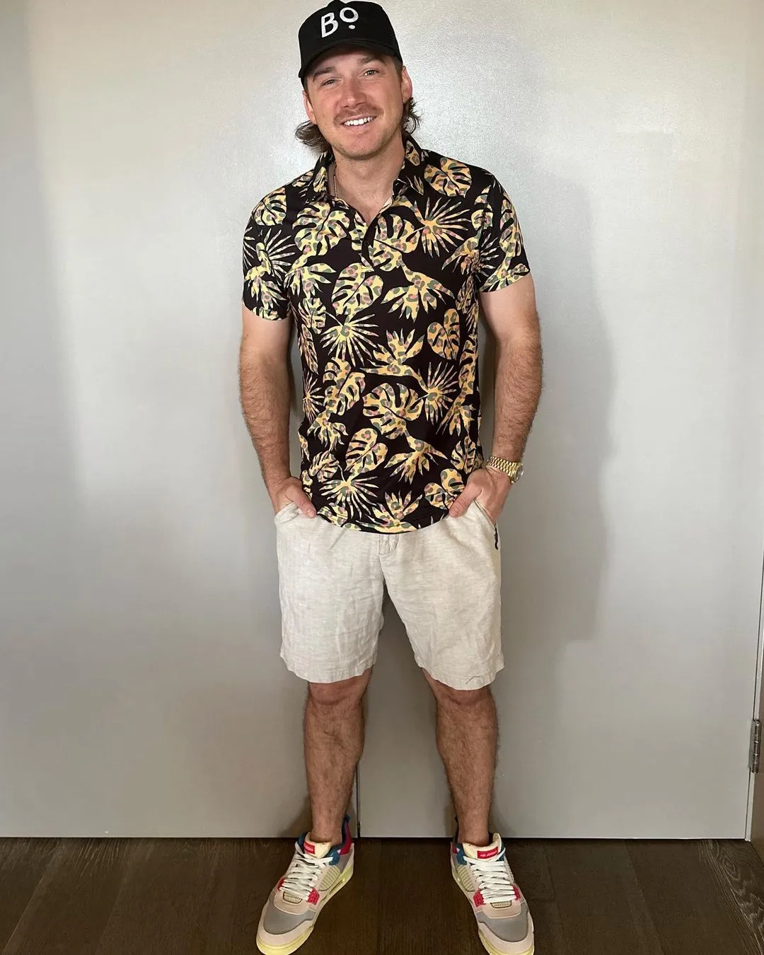 Morgan Wallen wearing a black cap, a black short-sleeved shirt with yellow and green leaf patterns, light-colored shorts, and multi-colored sneakers.