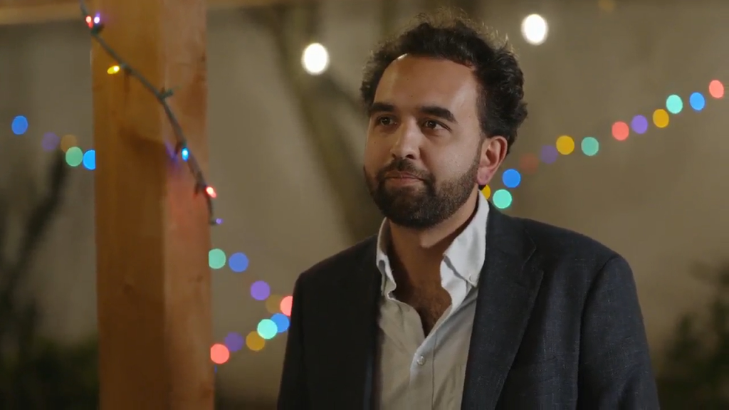 Man in a dark blazer and white shirt looking to the right, with blurred colorful string lights in the background.