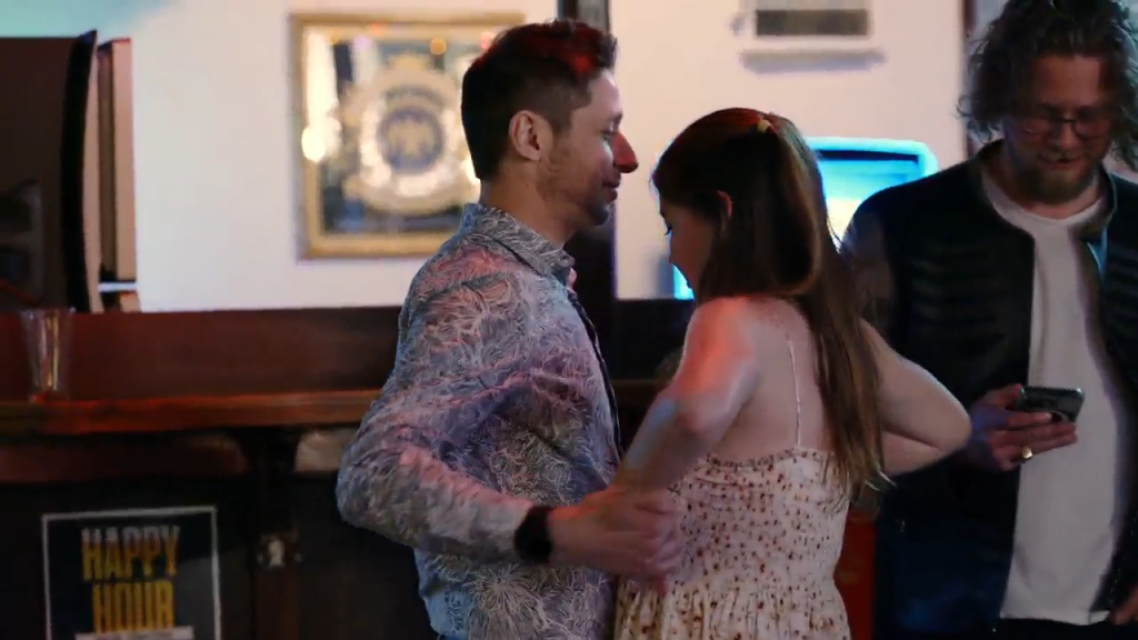 Catie and a man dancing at a bar while Josh watches from behind, looking at his phone.