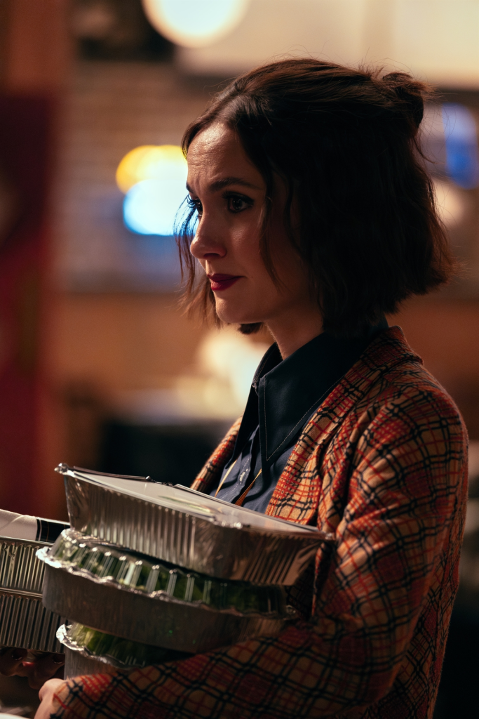 Woman in a plaid jacket carrying foil takeout containers.