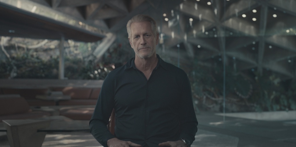 A middle-aged man with graying hair and beard, wearing a dark shirt, sitting in a modern room with large windows overlooking lush greenery.
