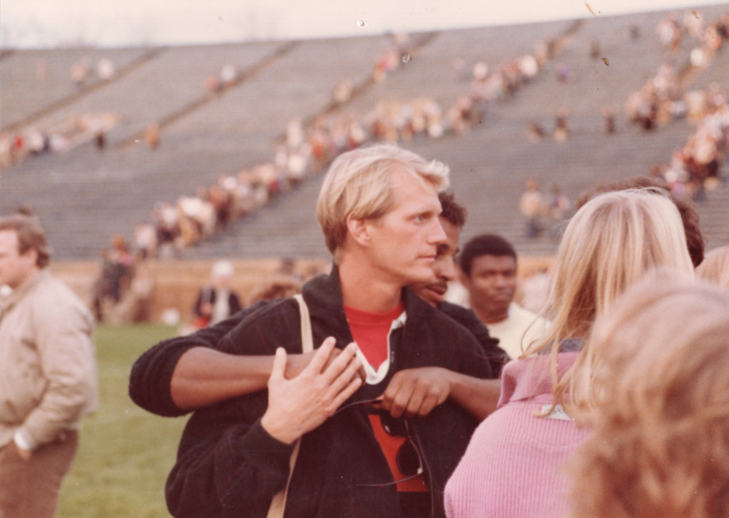Blonde man in a red shirt with arms around him, a stadium with spectators in the background.