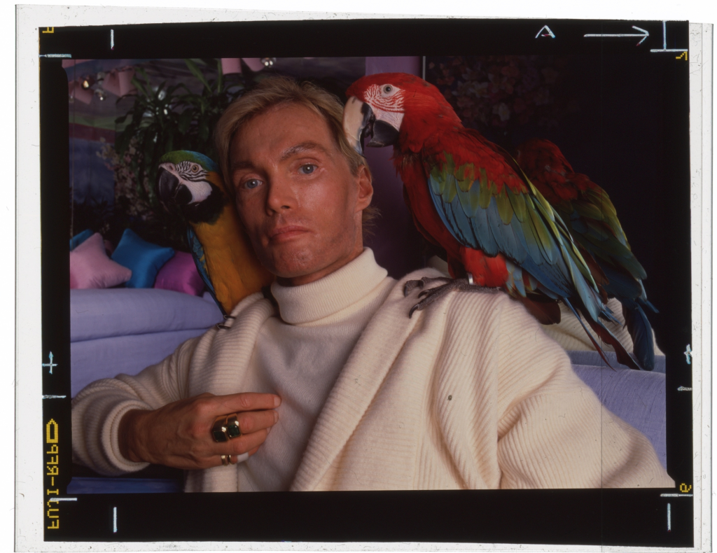 A man with a blue and gold macaw on his left shoulder and a red and green macaw on his right.