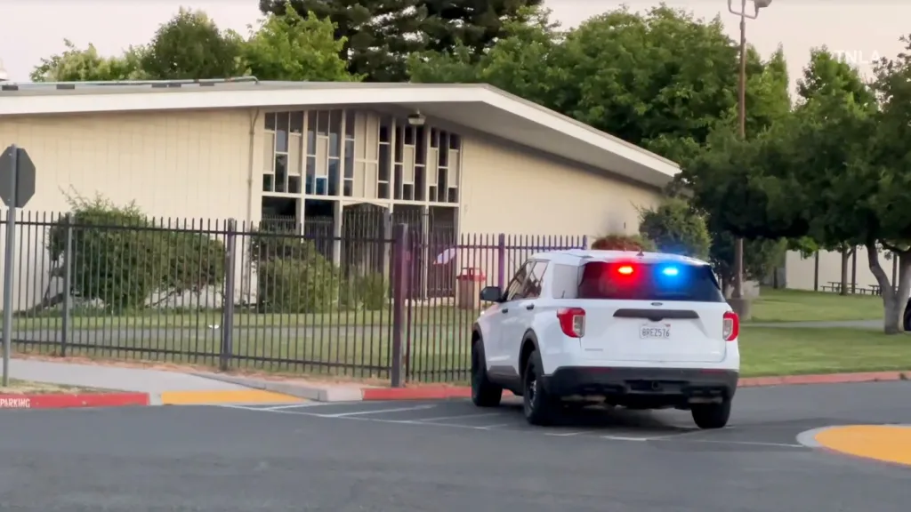 A white police SUV with flashing red and blue lights sits parked on the road next to a black fence, with a building and trees in the background.
