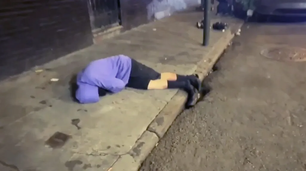 A person in a purple hoodie and black shorts lying face down on a sidewalk.