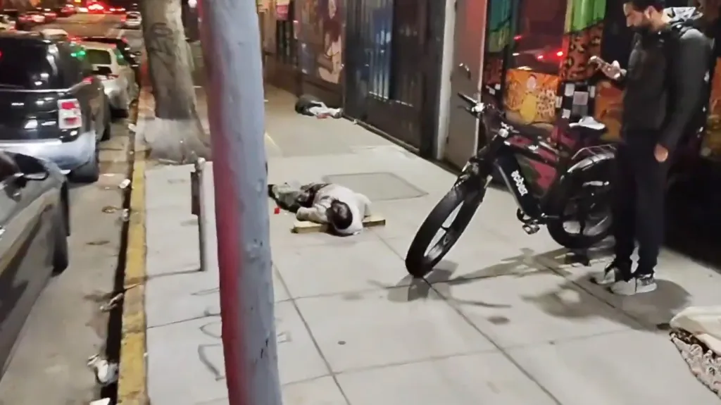 Three people are lying on a sidewalk, one of them next to a bicycle, with cars parked along the street.