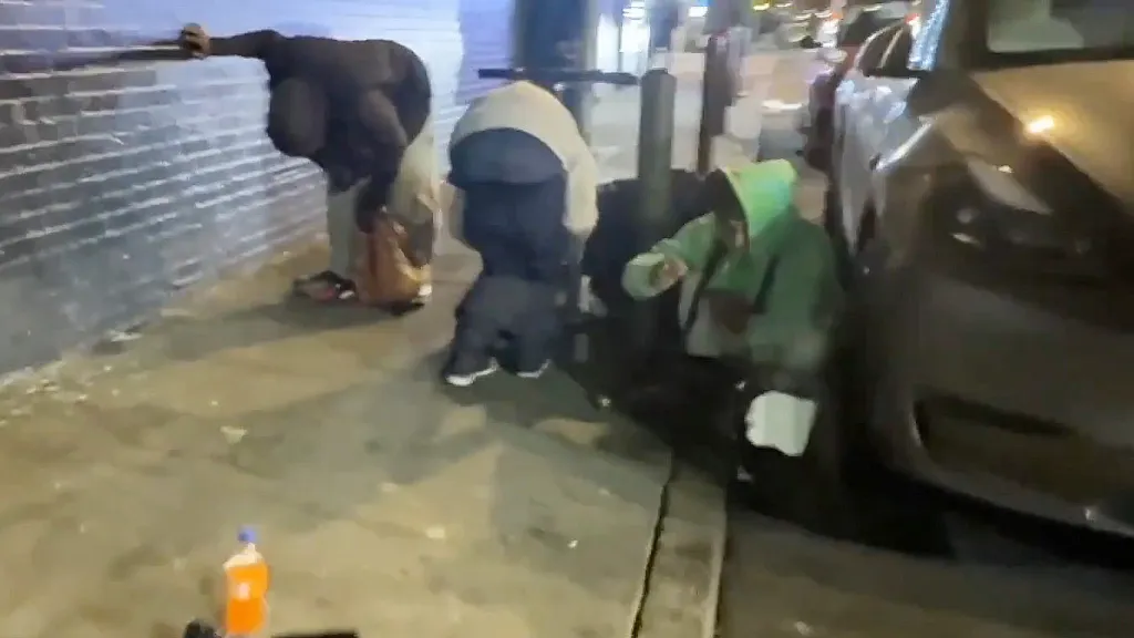 Three people crouched on a sidewalk at night, one near a brick wall, one near a pole, and one near a dark car, with an orange bottle on the ground.