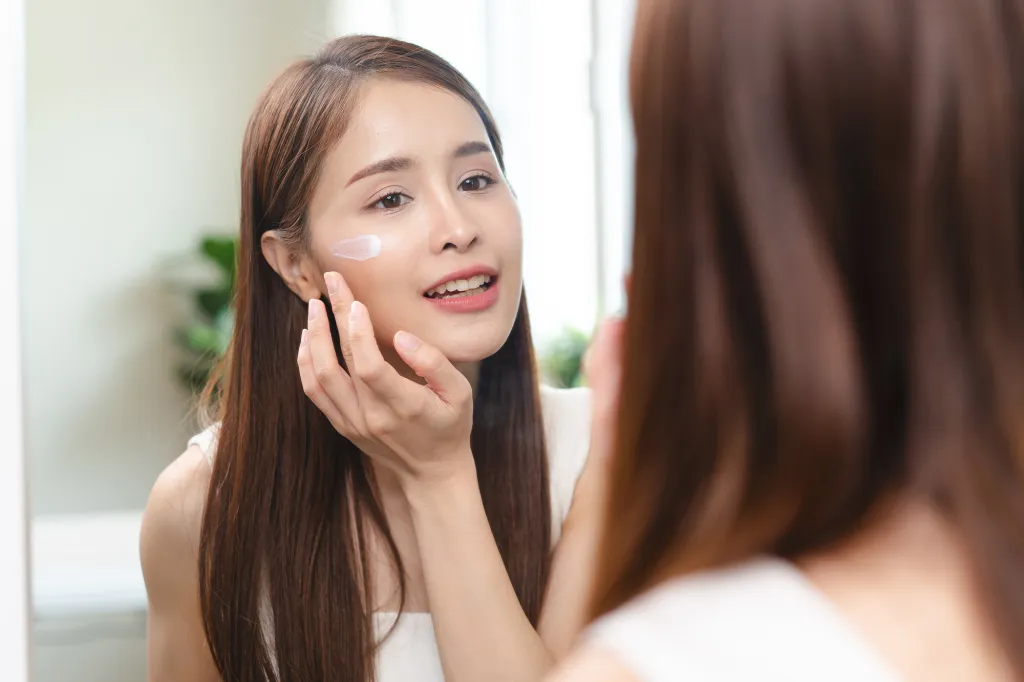 A woman looking in a mirror while applying cream to her face.