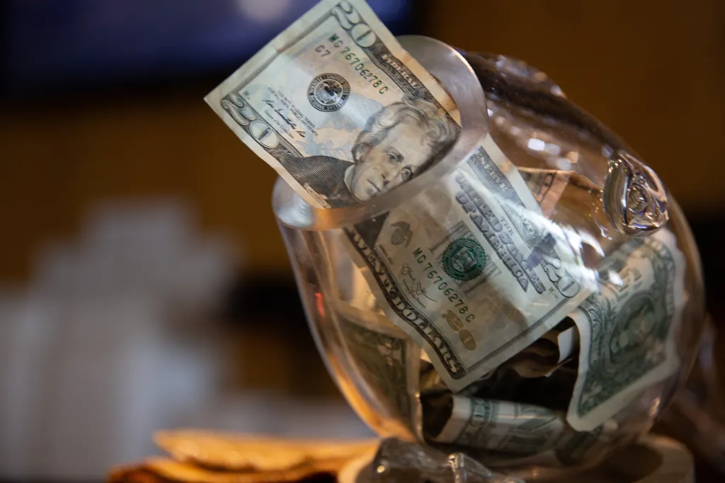 A glass tip jar containing several dollar bills, including a prominent $20 bill.