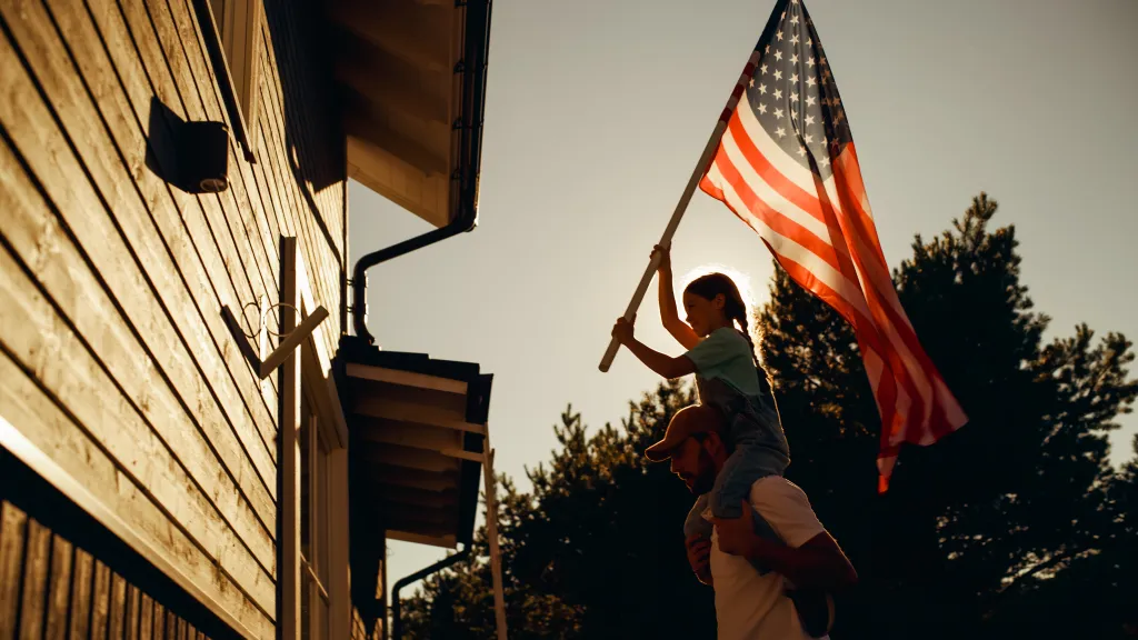 Father holding his daughter on his shoulders to help her raise an American flag at their house.