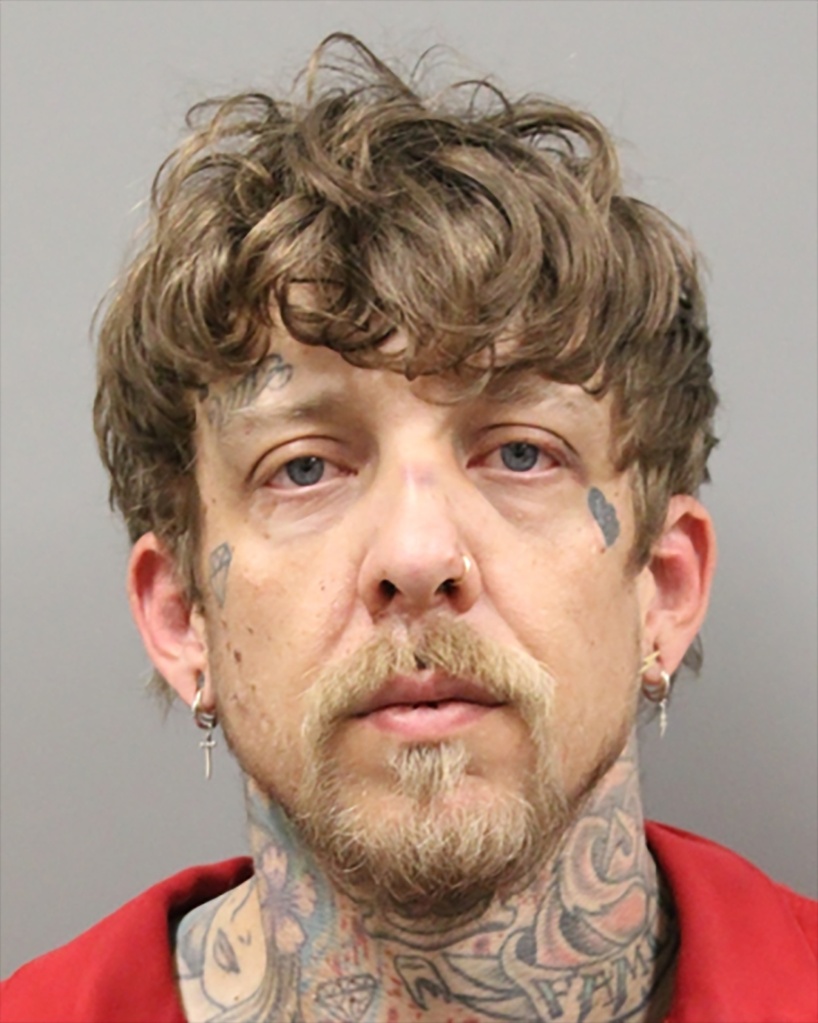 A mugshot of a man with shaggy brown hair, a patchy mustache and beard, and visible tattoos on his face and neck.