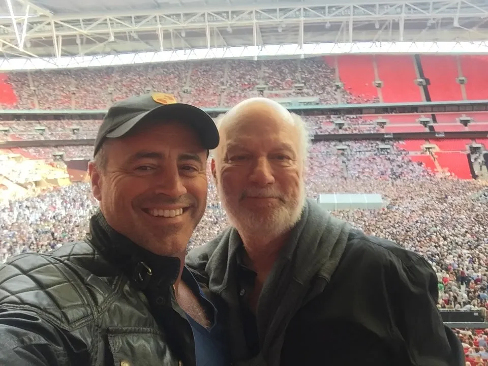 Matt LeBlanc and a man with a white beard take a selfie at a stadium.