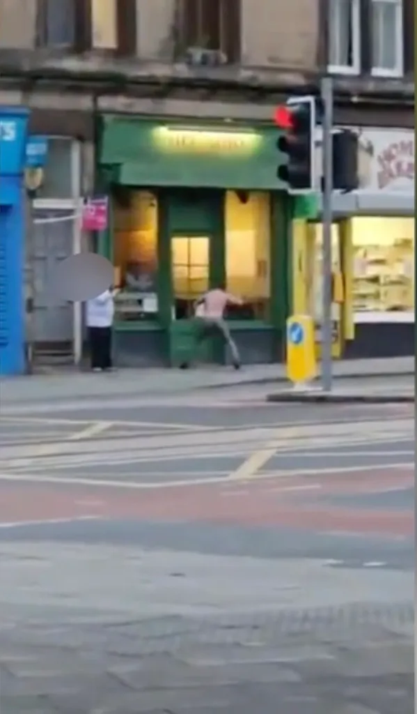 Man in a white shirt and a woman with an umbrella standing on the pavement, with a man running into a store in the background.