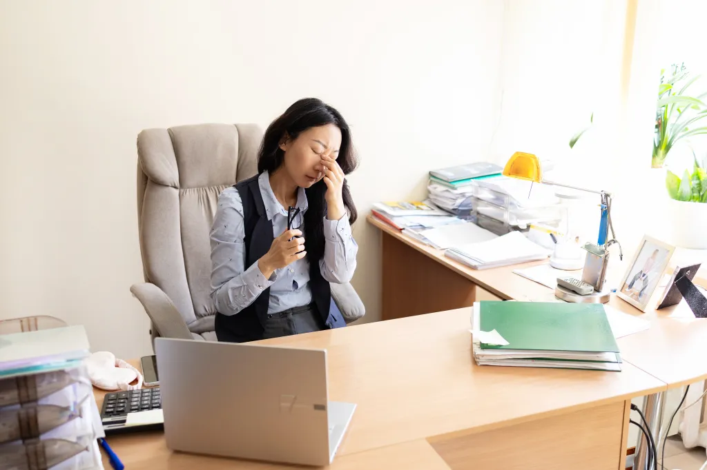 An Asian businesswoman rubs her eyes, looking stressed while sitting at a desk with a laptop and paperwork.