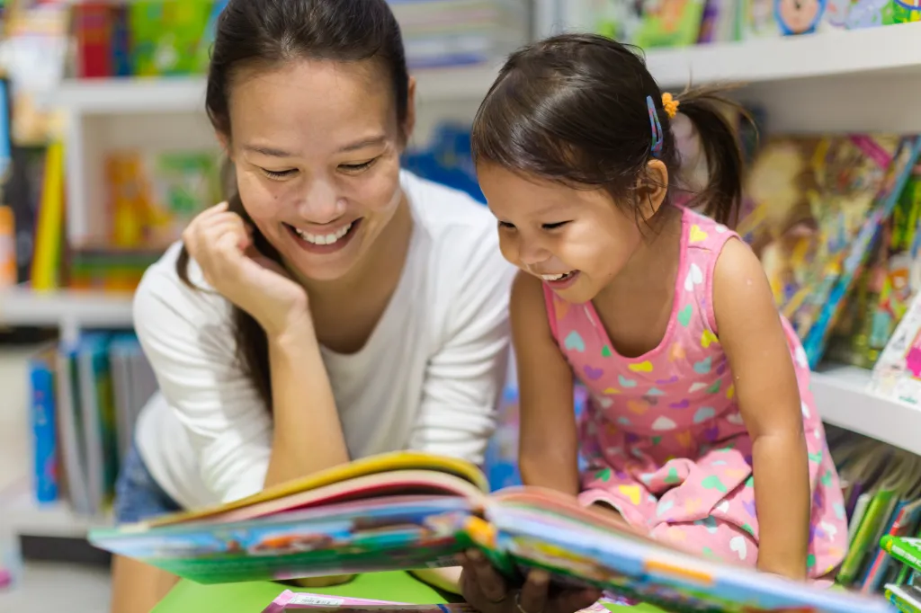 A mother and child laughing while reading a book in a library.