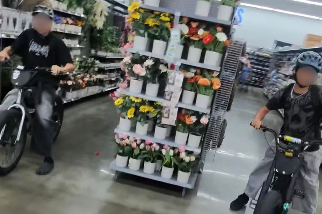 Two people riding mini bikes through a store aisle with artificial flowers.