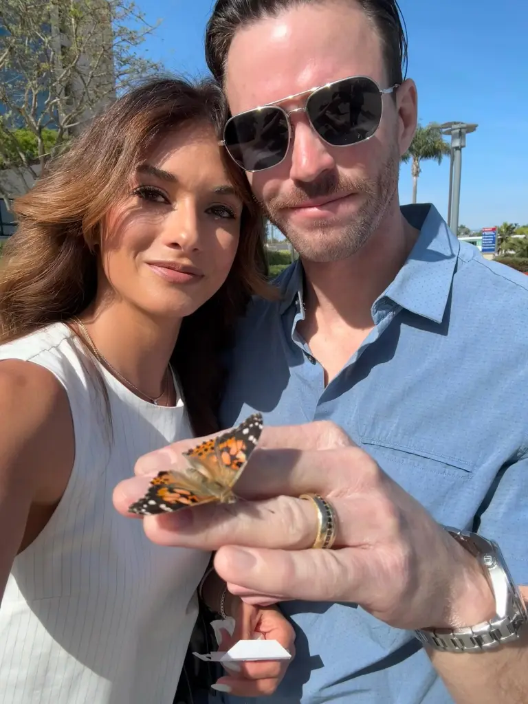 A woman in a white dress and a man in a blue shirt with sunglasses holding a butterfly on his hand.