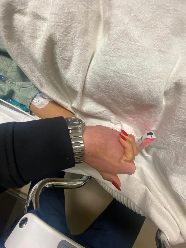 A man and woman holding hands in a hospital, the woman's hand has an IV drip and a pulse oximeter on her finger.