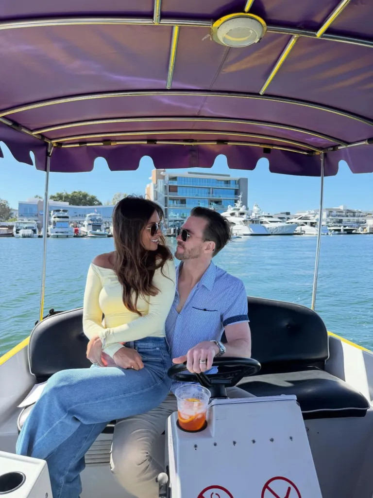 A couple sitting in a boat, looking at each other, with buildings and yachts in the background.