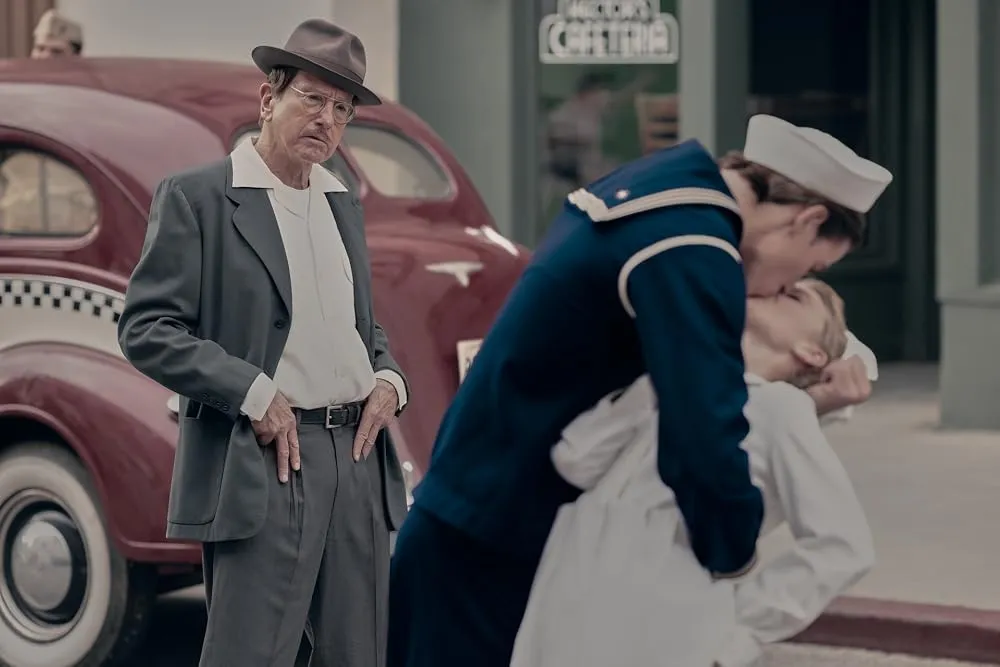 A man in a fedora and glasses looks displeased as a sailor in uniform kisses another person in the foreground.