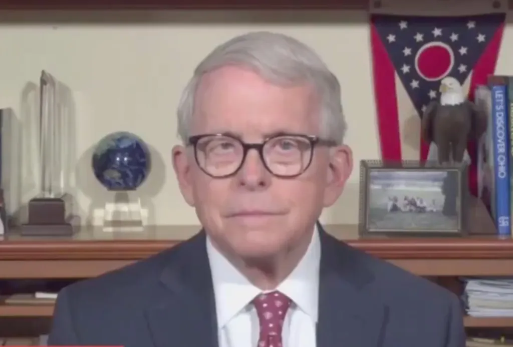 A man in glasses, a suit, and a red tie looks at the camera with an Ohio state flag and a bald eagle figurine behind him.