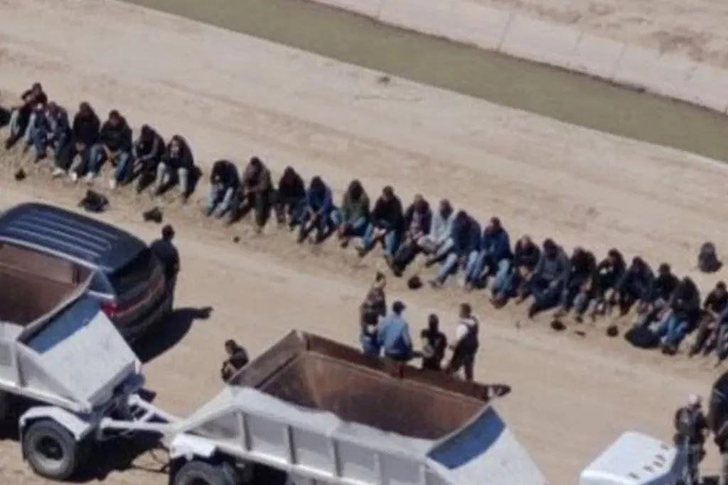 An aerial view of people sitting in a line along a dirt canal with law enforcement officers and vehicles nearby.